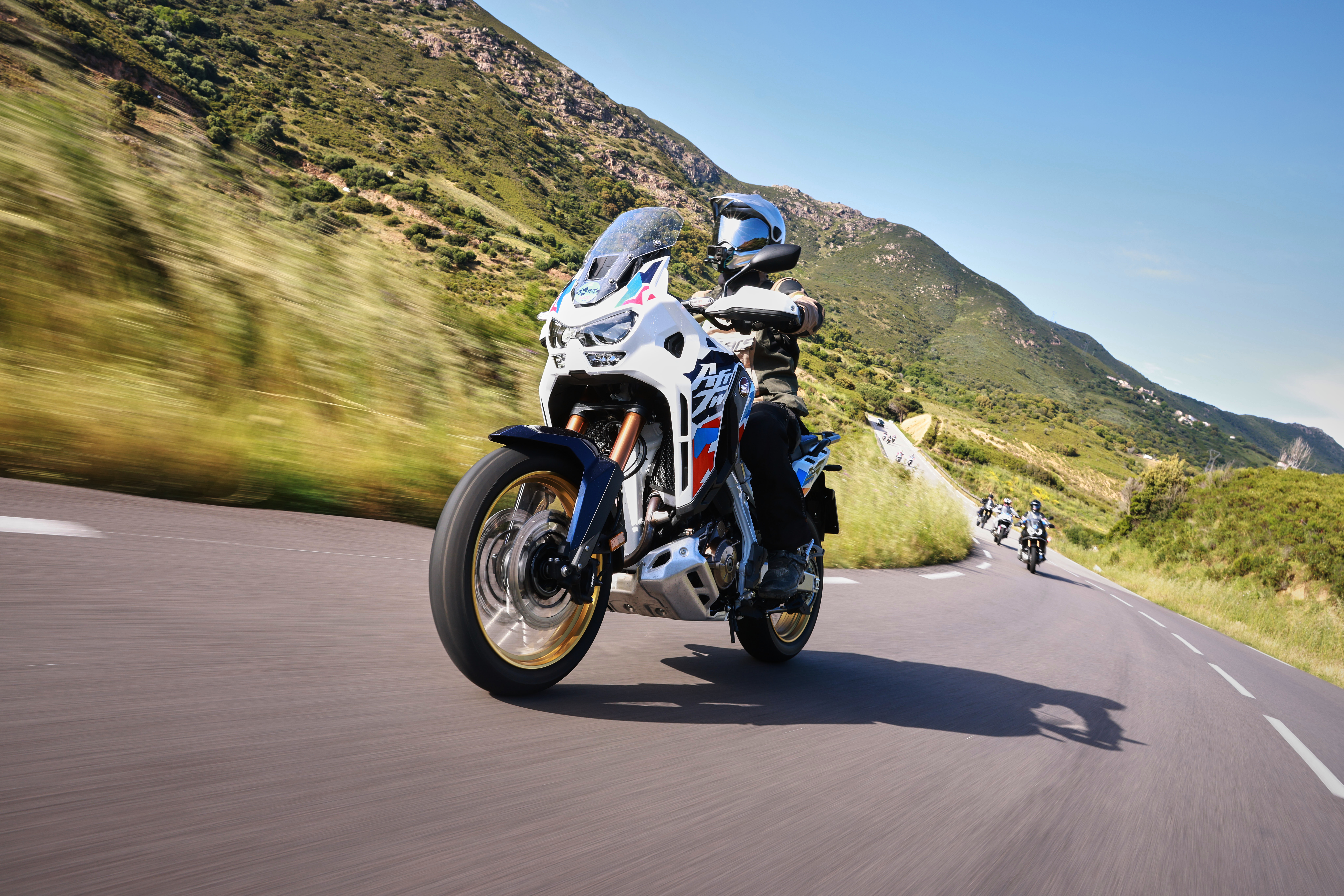 Una moto Honda Africa Twin in movimento su una strada tortuosa con montagne e colline sullo sfondo in una giornata di sole.